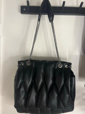 Svart quiltad handväska från Zara - Trendig svart handväska från Zara i quiltat mönster med silverfärgade kedjeremmar. Väskan har en mjuk, puffig design och är tillverkad i konstläder. Perfekt accessoar för att lyfta din outfit.