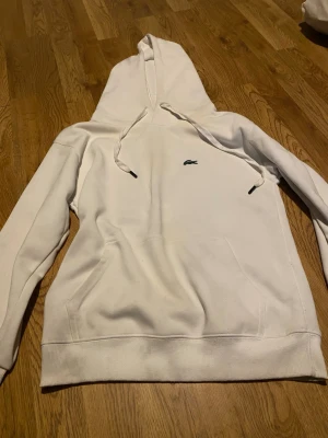 Vit hoodie från Lacoste  - Snygg vit hoodie från Lacoste med klassisk krokodil-logga på bröstet. Regular fit med stor huva och dragsko samt magficka framtill. Perfekt för en clean och stilren look. Mjuk bomullsblandning som känns skön mot huden.