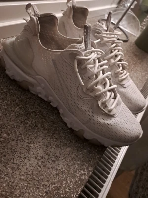 Vita Nike React - Snygga vita Nike React Element sneakers med modern design och unika detaljer. Skorna har en stickad ovandel i mesh med diskreta mönster, vita snören och en chunky sula med genomskinliga partier. Perfekta för dig som gillar streetstyle och sportig look.
