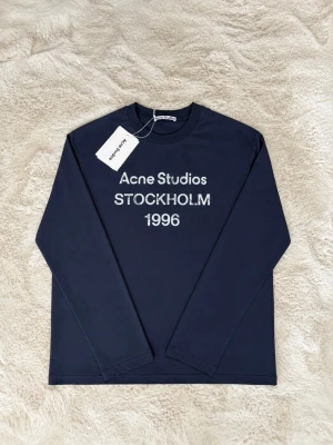 Acne Studios - Långärmad marinblå t-shirt från Acne Studios med vit text 'Acne Studios STOCKHOLM 1996' på bröstet. Klassisk rund hals och klassisk passform. Tröjan är aldrig använd och original lappen sitter kvar. Skriv om du vill ha mer bilder eller har frågor. Det är storlek S men sitter som M