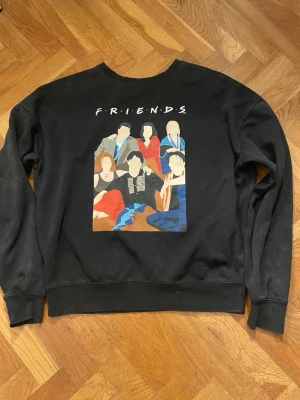 Svart Friends sweatshirt - Cool svart sweatshirt med tryck av Friends-gänget på framsidan och texten 'FRIENDS' ovanför. Tröjan har rund halsringning och ribbade muddar vid ärmslut och nederkant. Perfekt för dig som älskar tv-serien Friends och vill ha en avslappnad stil.