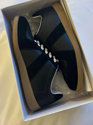 Maison Margiela svarta sneakers - Maison Margiela sneakers i svart läder med beige mockadetaljer och kontrasterande vita snören. Klassisk gummisula i ljusbrunt och ikonisk design. Perfekta för dig som gillar stilrena och trendiga skor med lyxig känsla.