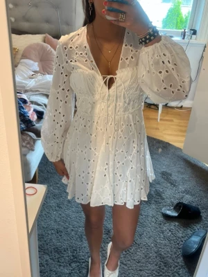 Student klänning  - Superfin vit kort klänning med broderade detaljer och hålmönster. Klänningen har långa ärmar, v-ringning och snörning framtill. Perfekt för sommarens alla tillfällen och ger en romantisk vibe. Passar perfekt som studentklänning