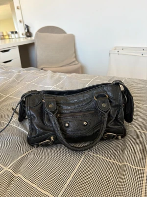 Svart handväska - Cool svart handväska i mjukt läder med klassiska nitar, dragkedja framtill och flätade handtag. Väskan har silvriga metalldetaljer och en unik, lite slouchy form. Perfekt för dig som gillar statement-accessoarer och ikonisk design.