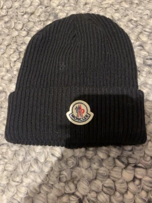 Svart Moncler ribbstickad mössa - Svart ribbstickad mössa från Moncler med uppvikt kant och klassisk Moncler-logga framtill. Tillverkad i mjukt material som håller dig varm och snygg under kalla dagar. Perfekt accessoar för en stilren look.