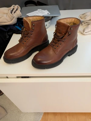 Bruna läderkängor med grov sula - Snygga bruna kängor i äkta läder med kraftig svart gummisula och snörning hela vägen upp. Klassisk modell med högt skaft och metallöljetter. Perfekta för dig som gillar en stilren och robust look.