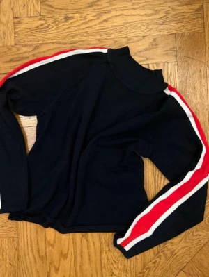 Svart stickad tröja med ränder - Svart stickad tröja med höghals och långa ärmar. Snygga röda och vita ränder längs båda ärmarna ger en sportig vibe. Perfekt för dig som gillar enkel men trendig stil.