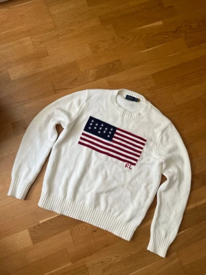 Vit stickad tröja Ralph Lauren - Snygg vit stickad tröja från Ralph Lauren med amerikansk flagga i rött, vitt och blått på bröstet samt RL-broderi. Tröjan har rund halsringning och ribbade muddar vid ärmslut och nederkant. Perfekt för en klassisk och avslappnad stil.