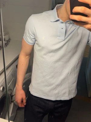 Ljusblå pikétröja från Polo Ralph Lauren - Snygg ljusblå pikétröja från Polo Ralph Lauren med klassisk krage och knappar framtill. Tröjan har korta ärmar och är tillverkad i mjuk bomull. Jag är 187cm och 80kg.