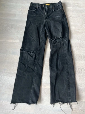 Svarta slitna jeans med raka ben - Coola svarta jeans med raka ben och slitna detaljer på knäna. Jeansen har råa, fransiga benslut och klassisk femficksmodell. Perfekta för en avslappnad streetstil. Materialet är denim i bomull.