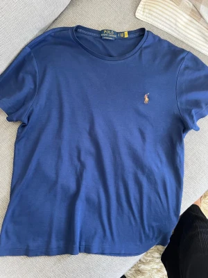 Blå t-shirt från Polo Ralph Lauren - Snygg blå t-shirt från Polo Ralph Lauren med klassisk rund hals och broderad logga på bröstet. T-shirten är enkel och stilren, perfekt till jeans eller shorts. Tillverkad i mjuk bomull för skön känsla hela dagen.