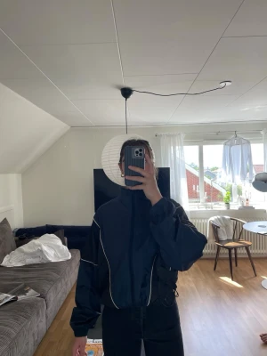 Mörkblå vindjacka med vita detaljer - Snygg mörkblå vindjacka med vita kontrastsömmar och dragkedja framtill. Jackan har hög krage, avslappnad passform och dragsko i midjan. 