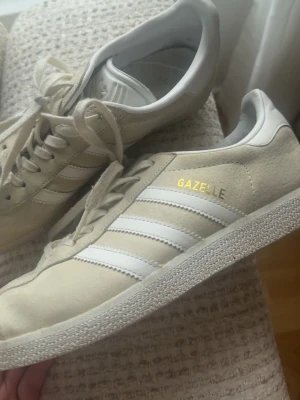 Adidas Gazelle beige sneakers - Säljer ett par klassiska Adidas Gazelle sneakers i beige mocka med vita detaljer och guldtext på sidan. Skorna har den ikoniska tre ränder-designen, vit sula och snörning. Perfekta för dig som gillar stilrena och tidlösa sneakers.