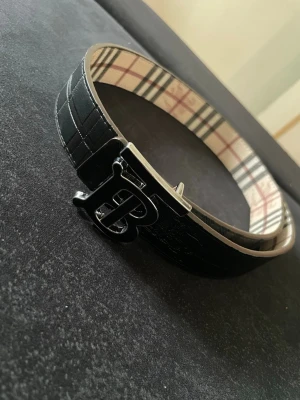 Svart Burberry bälte med rutigt foder - Stilrent svart bälte från Burberry med klassiskt rutigt foder på insidan och en stor metallspänne med Burberrys logotyp. Bältet är gjort i skinn och har en elegant, tidlös design som passar till många outfits.