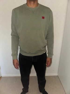 Les deux Olivgrön sweatshirt med röd patch - Olivgrön sweatshirt med rund hals och långa ärmar. Tröjan har en liten röd patch på bröstet och ribbade muddar vid ärmslut och nederkant. Perfekt för en avslappnad och stilren look.