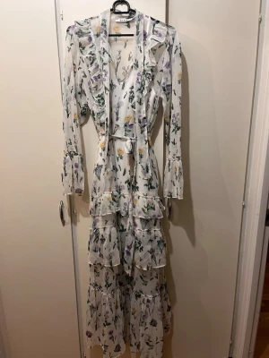 Lång maxi blommig vit klänning NA-KD strl 38 - Klänning från NA-KD i strl 38. Knytband i midja, volanger, långa ärmar. Till klänningen så medföljer tillhörande längre underklänning. Mycket fint skick. 