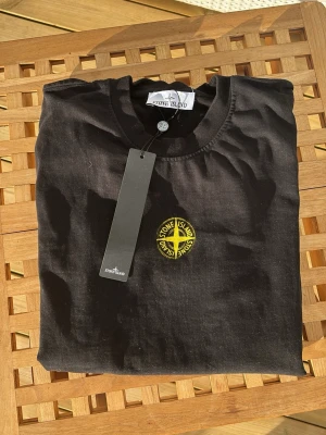 Stone Island t-shirt  - Svart t-shirt från Stone Island med klassisk rund hals och ikonisk gul logga broderad på bröstet. T-shirten är kortärmad och tillverkad i mjuk bomull, perfekt för en stilren och avslappnad look.