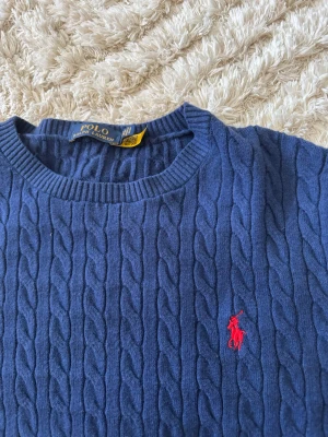 Blå kabelstickad tröja Polo Ralph Lauren - Kabelstickad blå tröja från Polo Ralph Lauren med rund halsringning och röd broderad logga på bröstet. Långa ärmar och ribbade muddar vid ärmslut och nederkant. Perfekt för en stilren och klassisk look.