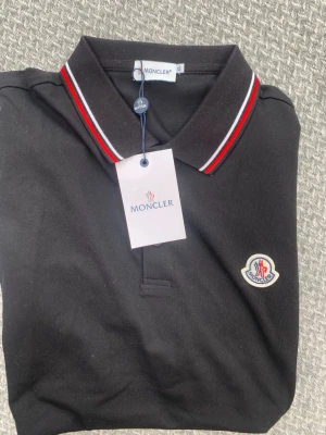 Svart pikétröja från Moncler - Svart pikétröja från Moncler med klassisk krage och röd-vit randig detalj på kragen. Tröjan har Moncler-logga broderad på bröstet och är tillverkad i bomull. Perfekt för en stilren och sportig look.