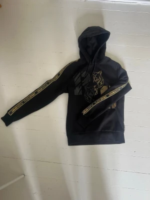 Svart hoodie från Plein Sport - Svart hoodie från Plein Sport med huva och dragsko. Framsidan har ett stort tryck. Guldiga band med logotyper längst ärmarna. Storlek S