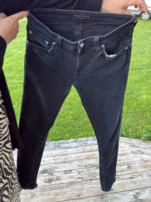 Svarta skinny jeans från Nudie - Säljer ett par svarta skinny jeans från Nudie med klassisk femficksdesign och diskreta slitningar på bakfickorna. Jeansen har låg midja och smal passform, perfekta för en trendig och avslappnad stil.