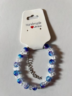Handgjort armband med blå pärlor - Ett handgjort armband med glittrande blå och genomskinliga pärlor. Armbandet har en justerbar silverfärgad kedja och är perfekt för att ge din stil en färgklick. Pärlorna är runda och skimrar i olika blå nyanser.