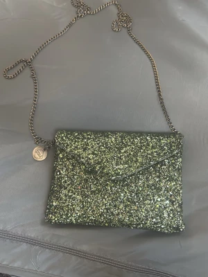 Grön glittrig kuvertväska med kedja - Säljer en grön glittrig kuvertväska från pipol’s bazaar med  kedjerem. Väskan har magnetlås och insida i ljus satin med ett litet innerfack. Perfekt för att addera lite extra bling till din outfit!