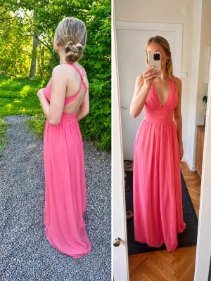 Rosa långklänning med öppen rygg - Snygg rosa långklänning med djup v-ringning och korsade axelband i ryggen. Klänningen har markerad midja och är tillverkad i ett lätt, flytande material som ger en elegant siluett. Perfekt för sommarens speciella tillfällen.