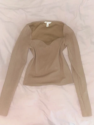 Beige ribbad långärmad topp H&M - Trendig beige ribbad topp från H&M med lång ärm och snygg sweetheart-urringning. Figurnära passform som framhäver midjan och ger en modern siluett. Perfekt att styla med jeans eller kjol för en clean look.