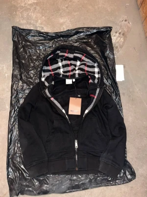 Svart Burberry Hoodie - Snygg svart hoodie från Burberry med dragkedja och klassiskt rutigt foder i huvan i svart, vitt och rött. Hoodien har fickor framtill och är perfekt för dig som vill ha en stilren och exklusiv look. Den är lite dammig, men den är helt ny går att borsta av.