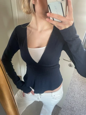 Svart omlottopp med v-ringning - Snygg svart omlottopp med v-ringning och lång ärm. Toppen har en ribbad struktur och sitter figurnära. Perfekt att styla med jeans eller kjol för en trendig look.