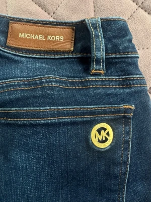 Mörkblå jeans från Michael Kors - Snygga mörkblå jeans från Michael Kors med klassisk femficksmodell och guldfärgad MK-detalj på fickan. Jeansen har kontrastsömmar och är tillverkade i ett mjukt denimtyg som ger en skön passform. Perfekta för dig som gillar stilrena och exklusiva detaljer. Väldigt snyggt till ett par klackar eller höga boots, liten i storlek.