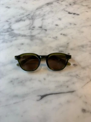 S&T 001 Green solglasögon  - Stig fram med stil och självförtroende i våra solglasögon 001. Med en elegant och modern design är dessa solglasögon det perfekta accessoaret för att komplettera din sommarlook. Tillverkade med högkvalitativa material och med omsorgsfullt utformade detaljer, erbjuder de inte bara en trendig estetik utan också överlägsen skydd mot solens strålar. Med sina mångsidiga och universellt smickrande form passar de alla ansiktsformer och ger en sofistikerad touch till varje outfit. 
