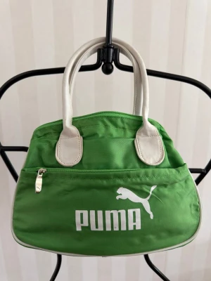 Grön handväska från Puma - Cool grön handväska från Puma med vita detaljer och handtag. Väskan har ett stort fack med dragkedja och en mindre ficka framtill. Tillverkad i slitstarkt tygmaterial med tydlig Puma-logga på framsidan. Perfekt för dig som gillar sportig stil.
