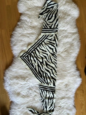 Zebramönstrad sjal i satin - Snygg zebramönstrad sjal i svart och vitt med breda ränder längs kanterna. Materialet känns glansigt och mjukt, troligen satin eller polyester. Perfekt att knyta runt halsen eller i håret för en trendig look.