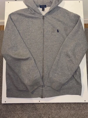 Grå hoodie från Polo Ralph Lauren - Klassisk grå hoodie med dragkedja från Polo Ralph Lauren. Tröjan har huva, två fickor framtill och den ikoniska mörkblå broderade loggan på bröstet. Tillverkad i mjuk bomullsblandning som är perfekt för en avslappnad stil. 170cm // 14/15år