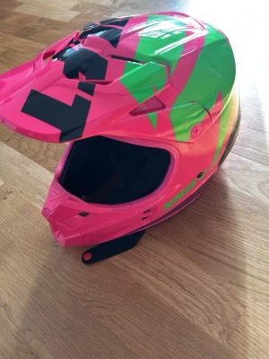 Färgglad crosshjälm Shift - Säljer en cool crosshjälm från Shift i stark rosa med gröna och svarta grafiska mönster. Hjälmen har skärm framtill och ventiler för bra luftflöde. Perfekt för dig som vill sticka ut på banan eller i skogen.