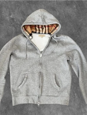 Grå hoodie från Burberry med dragkedja - Snygg grå hoodie från Burberry med dragkedja och klassiskt rutigt foder i huvan. Hoodien har två fickor framtill och justerbar dragsko i huvan. Perfekt för lager-på-lager och chill vardagsstil.🐧