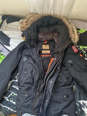 Svart parkas från Parajumpers - Snygg svart parkas från Parajumpers med fuskpäls på huvan och flera praktiska fickor. Jackan har orange detaljer, patch på ärmen och varm fodring. Perfekt för kalla dagar och riktigt streetig stil.