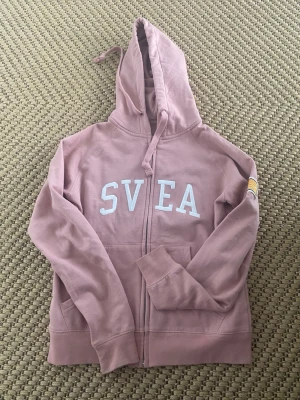 Rosa hoodie med dragkedja från SVEA - Rosa hoodie från SVEA med vit logga broderad på bröstet och en patch med amerikansk flagga på ärmen. Hoodien har dragkedja framtill, huva med dragsko och ribbade muddar. Tillverkad i mjukt bomullsmaterial, perfekt för en avslappnad stil.💞