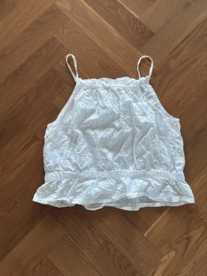 Vit linnetopp  - Söt vit linnetopp med tunna axelband och broderade blommor. Toppen har resår i midjan och en volangkant nedtill som ger en romantisk känsla. Perfekt för varma dagar och enkel att matcha med jeans eller kjol.