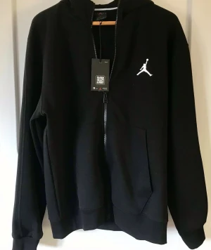 Svart hoodie från Jordan med dragkedja - Snygg svart hoodie från Jordan med dragkedja framtill och vit Jumpman-logga på bröstet. Klassisk huva och stora fickor framtill. Tillverkad i mjukt material som passar perfekt till streetwear-stilen.