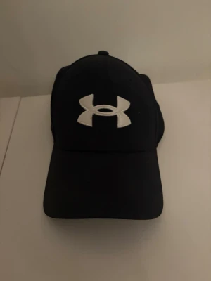 Svart keps från Under Armour - Snygg svart keps från Under Armour med det klassiska vita logotrycket framtill och broderad text bak. Kepsen har böjd skärm och är tillverkad i ett mjukt, slitstarkt material som passar perfekt till streetwear eller sportiga outfits.