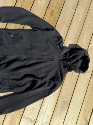 Svart hoodie från BOSS med dragkedja - Säljer en svart hoodie från BOSS med dragkedja framtill och två fickor med knappstängning. Hoodien har en klassisk huva och en diskret BOSS-logga på ärmen. Tillverkad i mjukt bomullsmaterial som känns skönt mot huden. Perfekt för en avslappnad och stilren look. Har ett hål i sig