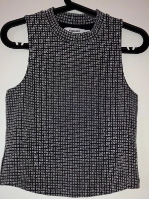 Glittrig ärmlös topp från Kappahl - Säljer en snygg ärmlös topp från Kappahl med glittrigt rutigt mönster i svart och silver. Toppen har en hög rund hals och är croppad i modellen. Perfekt för dig som vill sticka ut med lite extra shimmer.
