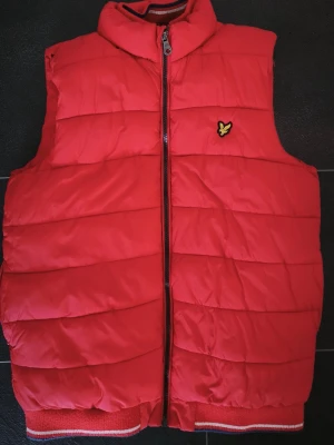 Röd dunväst från Lyle & Scott - Riktigt snygg röd dunväst från Lyle & Scott med quiltad design och dragkedja framtill. Västen har hög krage och ett broderat gult logomärke på bröstet. Ribbad nederkant med vita och grå detaljer. Perfekt för lager-på-lager under kalla dagar.