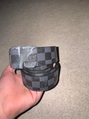 Svart och grått rutigt bälte - Snyggt bälte med svart och grått rutmönster och metallspänne. Bältet har en klassisk design och passar perfekt till jeans eller byxor. Materialet känns slitstarkt och ger en exklusiv känsla.
