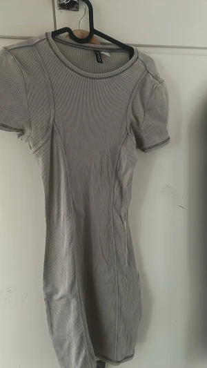 Ribbad kortärmad klänning H&M - Säljer en ribbad, kortärmad klänning från H&M i ljusgrått med smala ränder. Klänningen har en kroppsnära passform och rund halsringning. Perfekt för en enkel och stilren look.