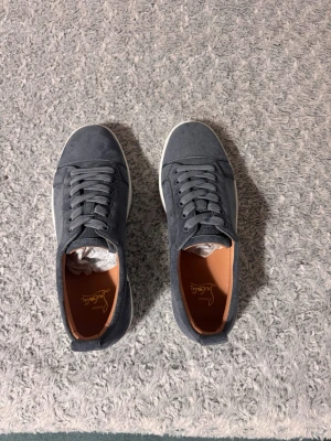 Ljus blåa Louboutin sneakers, storlek 43 - Snygga grå sneakers från Christian Louboutin i mjuk mocka med klassisk röd sula och vita detaljer. Lågt skaft, rund tå och matchande grå skosnören. Insidan är fodrad i brunt läder för extra komfort. Perfekta för dig som vill sticka ut med stil.