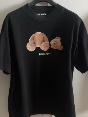 Svart Palm Angels t-shirt med nalle - Cool svart t-shirt från Palm Angels med ett unikt tryck av en nalle med avtagbart huvud på bröstet. T-shirten har klassisk rund hals och korta ärmar. Perfekt för dig som gillar streetwear och vill sticka ut med en edgy design.